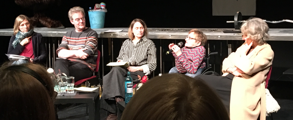 Vier Frauen und ein Mann sitzen nebeneinander auf einer Bühne. Eine Frau sitzt in einem Rolllstuhl und spricht in ein Mikrofon, die anderen schauen sie an.