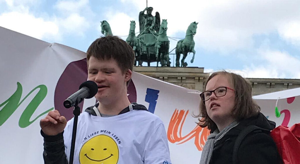 Ein junger Mann mit dunklen Haaren spricht in ein Mikrofon. Neben ihm steht eine unge Frau mit mittellangen Haaren und Brille. Beide haben Down-Syndrom. Im Hintergrund sieht man das Brandenburger Tor.