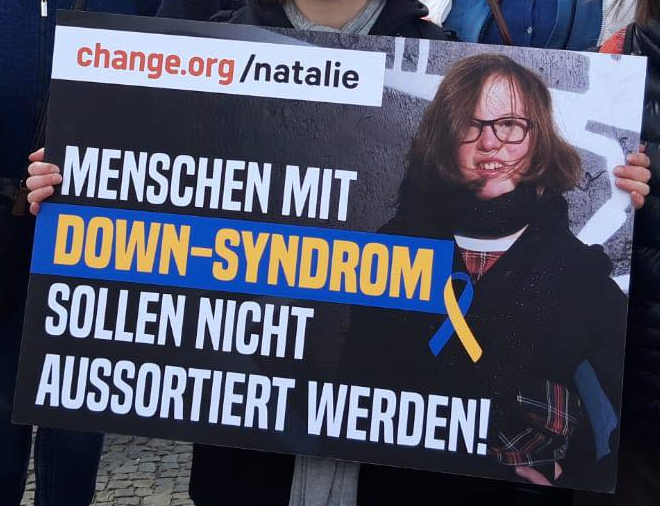 Plakat mit der Aufschrift: change.org/natalie - Menschen mit Down-Syndrom sollen nicht aussortiert werden! Hinter der Schrift liegt ein Foto von einer jungen Frau mit mittellangen dunkelblonden Haaren, dunkler Brille und Down-Syndrom.