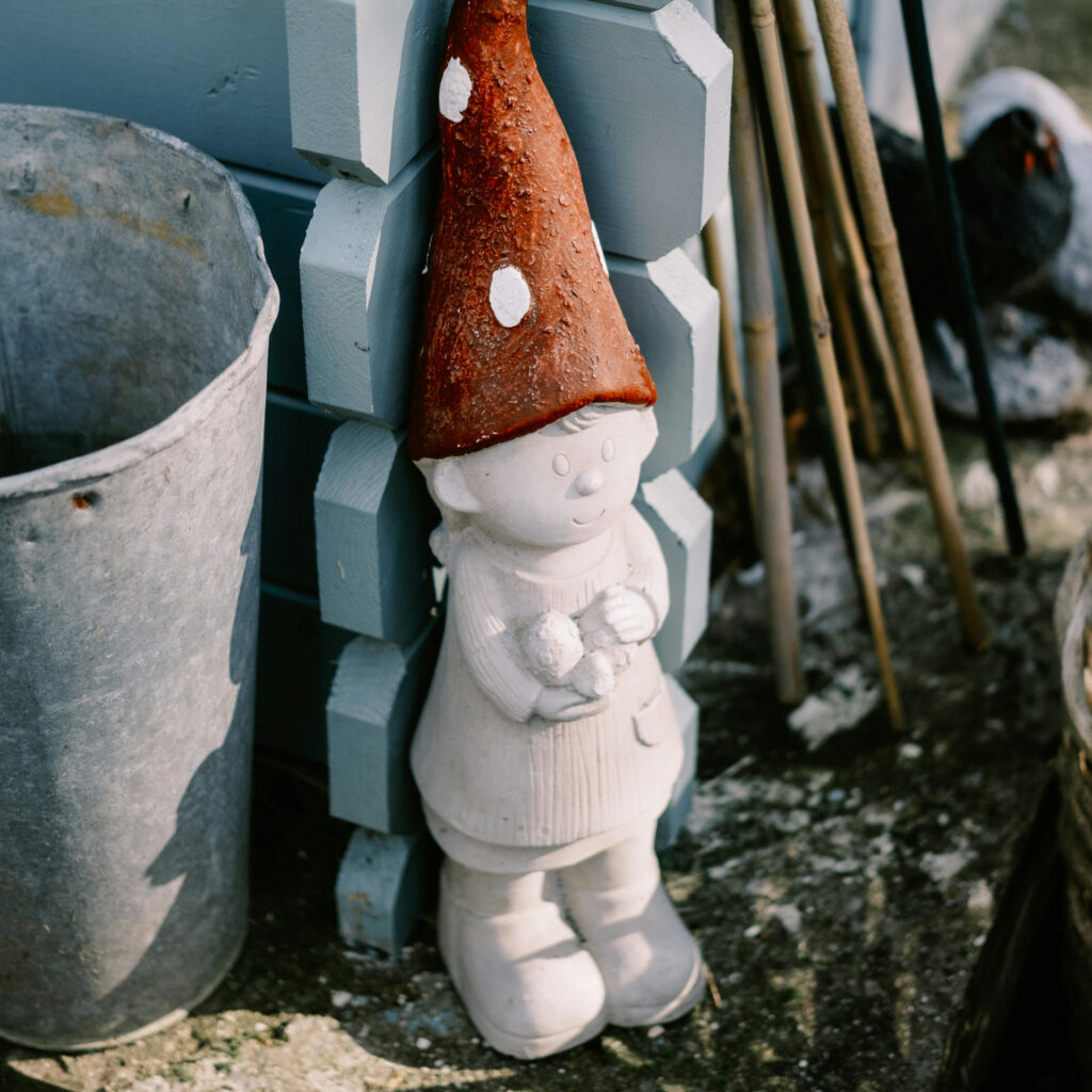 Imagebild zum Beitrag: Ein weißer kindlicher Gartenzwerg mit einer roten Zwergenmütze mit weißen Punkten steht mit dem Rücken zur Wand in einer Ecke im Garten.