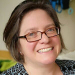 Porträt von Inga Oppenhausen. Sie hat kinnlange braune Haare, trägte eine Brille und eine gemusterte Bluse und lächelt in die Kamera.